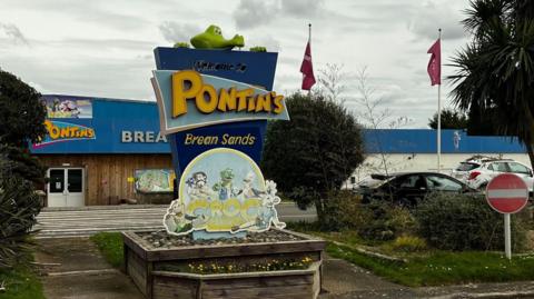 The exterior of Pontins Brean Sands, taken on a grey overcast day. It has a blue and yellow sign with a crocodile peering over the top, and there are two flag posts in the background. There is a small car park in front of the low single-story building. 