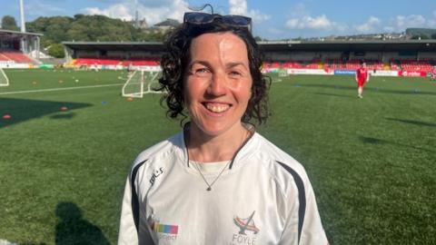 Meabh O'Neill stands in the middle of a football pitch. An empty stand filled with red seats is in the background.  A lone figure dressed in a red kit walks towards her. The woman is standing in front of a set of goals and is wearing a white football shirt with the words Foyle pride written on it.