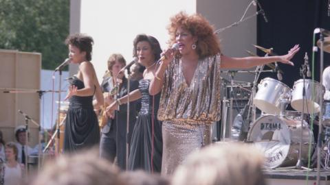A mid-shot of three women performing on an open-air stage in bright sunshine. One woman to the right of frame is slightly closer to the camera, wearing  a glamorous, silver-gold sparkling dress, and singing into a handheld microphone held in her right hand. She is about 40 and has slightly ginger-tinged dark hair in a large bouffant. To the left of frame and slightly behind on stage are two other singers in black dresses and with black hair, both slightly younger-looking, singing into microphones on stands.