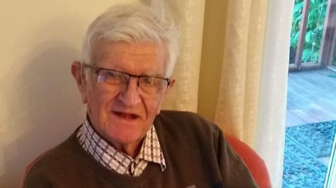 An old man with grey hair and glasses sits in a chair and smiles at the camera.