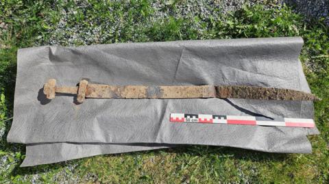 an ancient rusty viking sword laid out on a mat next to a ruler