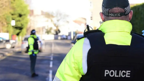 Two police officers on a road. One is facing away from the camera wearing a hat and the other is blurred in the background. 