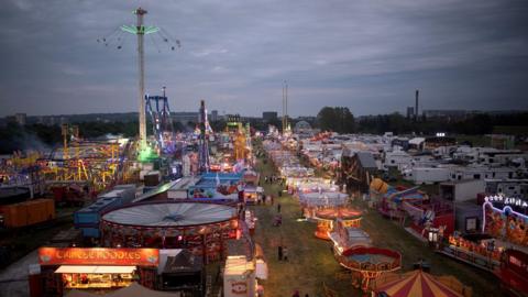 The Hoppings at dusk with rows of light up rides, the tallest being a tower with swings flying around the top. There are rows of white caravans to the right and a red Chinese noodles stall in the foreground.