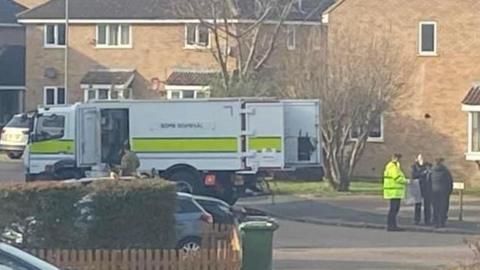 A housing estate with a white bomb disposal lorry with a yellow stripe and writing saying bomb disposal in black on the left. On the right is a police office in a yellow high-vis jacket talking to two dark-clad people 