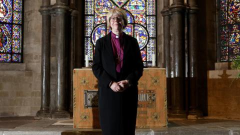 A woman wearing a purple religious tunic and a black coat stood in front of a series of stained glass windows and a large wooden table, which appears old. She is looking at the camera and is in a gothic cathedral building.