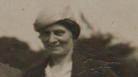 Louisa Dunne in a black and white photograph. She is wearing a hat and a brown jacket with a white shirt. 