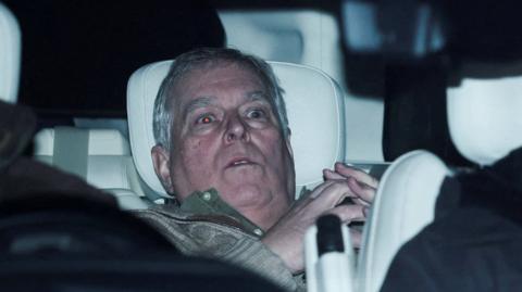 Andrew Mountbatten-Windsor is slumped in the back seat of a car with a white headrest. He looks shocked and has grey hair. He is wearing a green jumper and his hands are steepled together. His eyes are wide and the flash has turned them red.