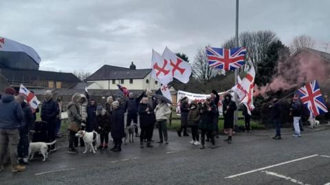 Protesters with St George's flags and Union Jacks, banners and flares gather in coats with dogs on the road outside the house.