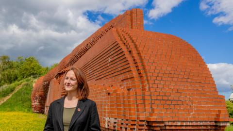 Lola McEvoy, Darlington MP, stands smiling next to the Darlington brick train. She is wearing a green top and a black suit jacket.