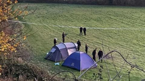 Aerial view of two blue tents in a field with a number of police officers standing around and blue police tape stretched across