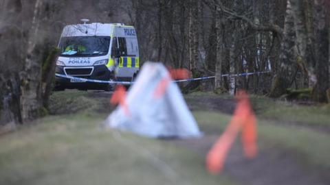 A police van is parked on a path in a wooded area. A small tent covering evidence is in the foreground.