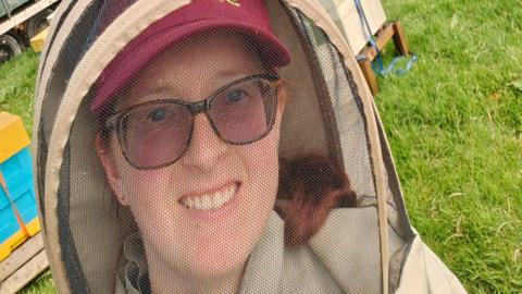 Christine with her beekeepers suit on standing in front of yellow and blue coloured bee hive boxes. Her hair is tied back and she is wearing glasses and a baseball cap.