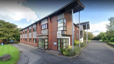 A brown brick building with brown framed windows and a concrete path around the corner of it