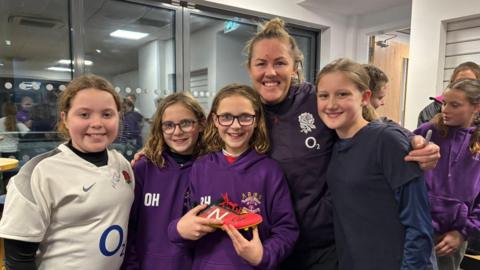 Marlie stands with a a group of four young girls who are part of the rugby team. Marlie wears an England training top with the rose emblem on it, and one of the girls is holding a signed boot. 