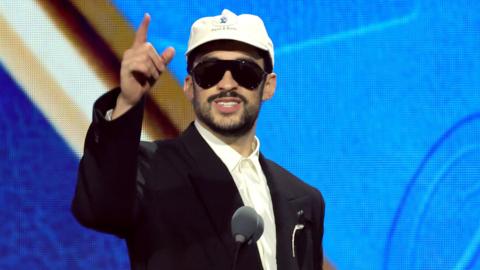 Bad Bunny accepting a Grammy award. He is wearing a blacksuit with shite shirt, a apir of sunglasses and a white cap. 
