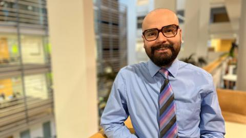 A man wearing a blue shirt with a stripy pink tie. He has large glasses and a brown beard.