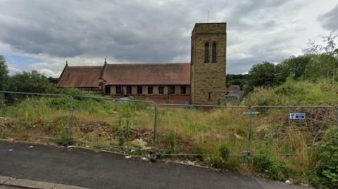 A metal fence surrounds a patch of overgrown land in front of a church