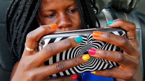 A woman with braids and multi-coloured nail varnish hold ups a mobile phone encased in a zebra design cover