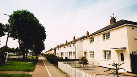 A row of lightly-coloured rendered houses. The one in the foreground has a white picket fence, with others having hedges along the front. There is a green space to the front of the properties.