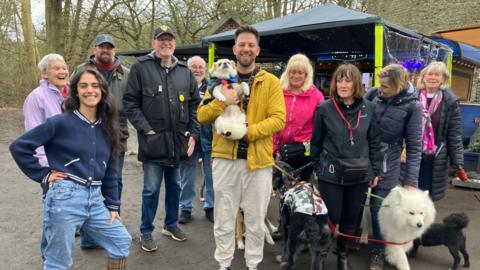 A group of people stand outside, in a park with their dogs. The coffee stand is in the background, with an awning for people to sit underneath.