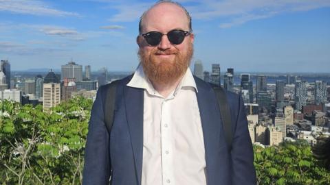A man wearing sunglasses and smiling in a blue suit jacket and white shirt, with skyscrapers and green foliage visible in the background