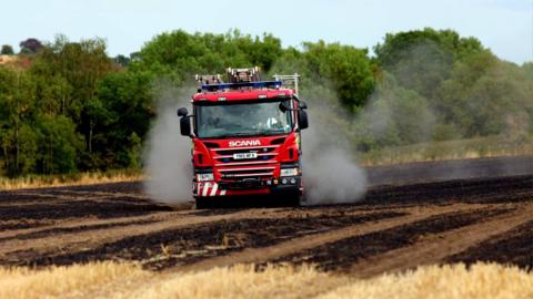 A fire engine driving through a burned cornfield.

There is smoke visible in the background.