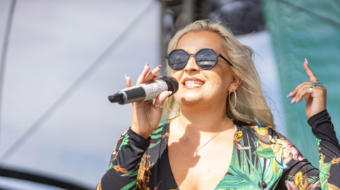 Sheddy Maria wearing a black floral shirt, black sunglasses, holding a black microphone