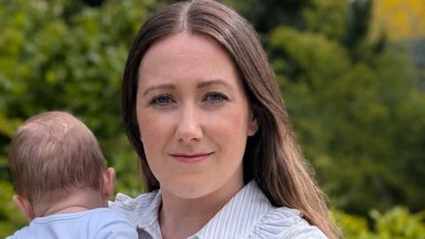 Rachael Bews smiles while holding her baby, whose face is turned away from the camera. They are in front of green trees