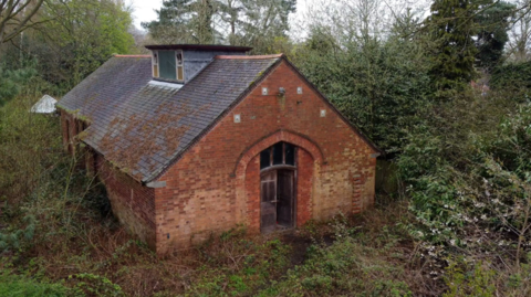 A red brick building with a large wooden door, and a pitched tiled roof with windows at the top. It is surrounded by trees and bushes