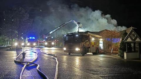 Fire engines parked in front of an on fire building, with smoke rising into the sky