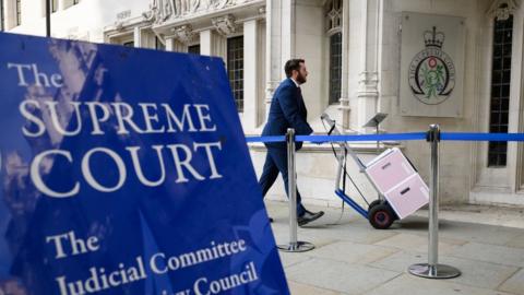 A man with boxes of documents on a trolley walks into the Supreme Court, with a blue sign in the foreground 