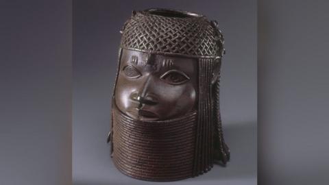 Commemorative head of the Oba, or king. It is a dark brown metal sculpture showing a face with a chain-mail helmet on its head and a metal collar up to its chin.