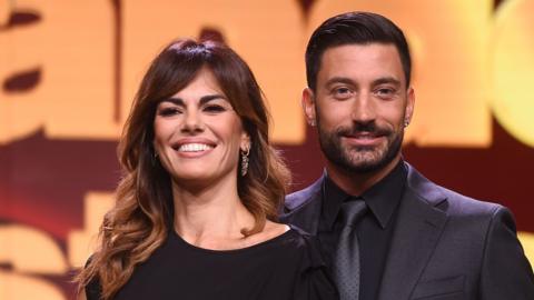 Giovanni Pernice holds arm around the waist of his partner Bianca infront of a dark red screen with the words Ballando Con Le Stelle on it. She is wearing a black dress and he a full black shirt, suit and tie. They are both smiling