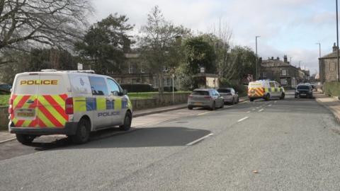 A public street with two police vans and parked cars. A bus stop, trees and other street furniture can be seen.