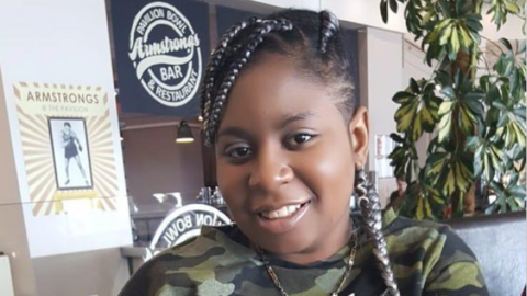 Edwige Nsilu sitting in front of a house plant at a bar. She has black hair, with braids that have white highlights in them. She is wearing a necklace and a green camoflague top and is smiling at the camera.