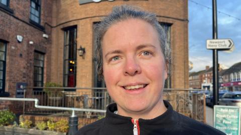 Gemma looking into the camera, her hair is tied back and she's wearing a black zip up jumper. Behind her is Bod Cafe and Bar, it's a brown brick building with some pub tables outside. In the far background (on the right) you can see a bit of Newport's high street.