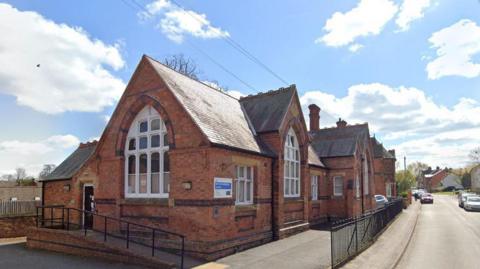 A small, single storey, red brick, Victorian school building by the side of a quiet village road, with a small number of houses visible at the end of the road.