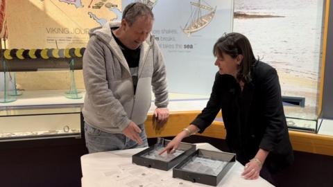 David O'Hare and Allison Fox looking at the silver coins, which are in plastic cases in two black drawers on a table in the Viking Gallery in the Manx Museum. They are standing in front of panels displaying information about the Viking age and glass display cases holding other Viking treasure.