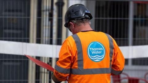 A Thames Water employee wearing a hi-vis uniform reads a document inside a red folder while on a site
