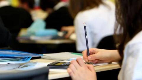 A close up of an anonymous female pupil's hands holding a pencil with a calculator on the desk with other pupils facing away from the camera in the background