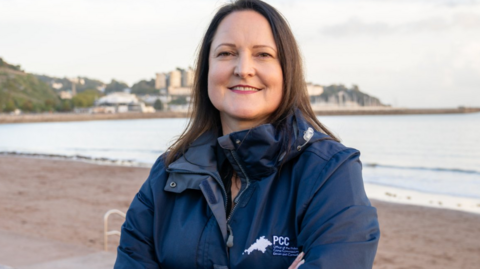A woman with shoulder length dark hair smiles at the camera as she wears a dark blue jacket with a logo and words on it with a seafront behind her.