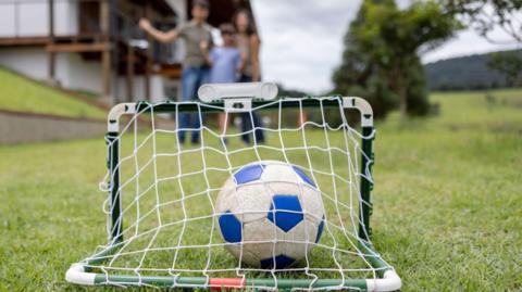 Foreground, a football sits inside a miniature goal. Behind we see two children celebrating