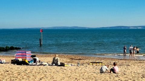 A beach scene with some people sat on the sand and a few others venturing out into the water. The blue sky is clear with hills in the background beyond the water.