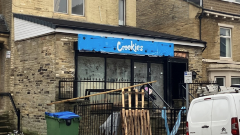 Shows a shop in a residential street. It has a blue sight with 'Crookies' written on it. A white van is parked outside it. 