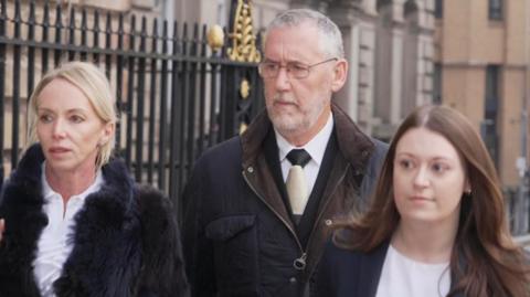 A man with grey hair and a suit walks along with two women