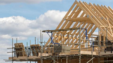 A wooden frame to a houses roof sits below a blue sky with partial cloud. There is scaffolding in front on the roof. 