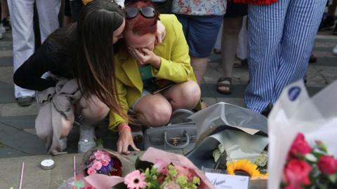 A woman in a yellow jacket and green dress is consoled by a younger woman in a black jumper as they place flowers among numerous floral tributes in Southport