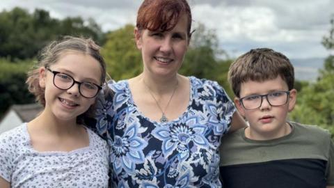 Picture of mum smiling, with her son and daughter on either side of her. The mum has short red hair and wears a dark blue and light blue floral dress. She wears a pendant on a chain necklace. Her daughter is on her right, wearing black-rimmed glasses and a white dress with green spots on it. Her son, to her left, wears dark blue-rimmed glasses and a green top with a broad black horizontal stripe.