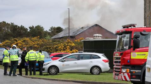 Smoke rises from a hole in the roof of a low, red brick building. The building is obscured by an area of gorse and low circular metal tank. A group of emergency services workers stand in a huddle next to a silver fire service car. There is also a fire engine parked nearby.