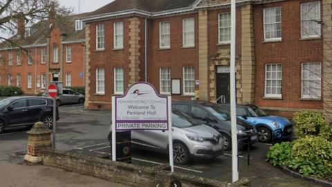 A two-storey building with brown bricks and beige edging. A sign saying welcome to Fenland Hall. Private parking is in front.